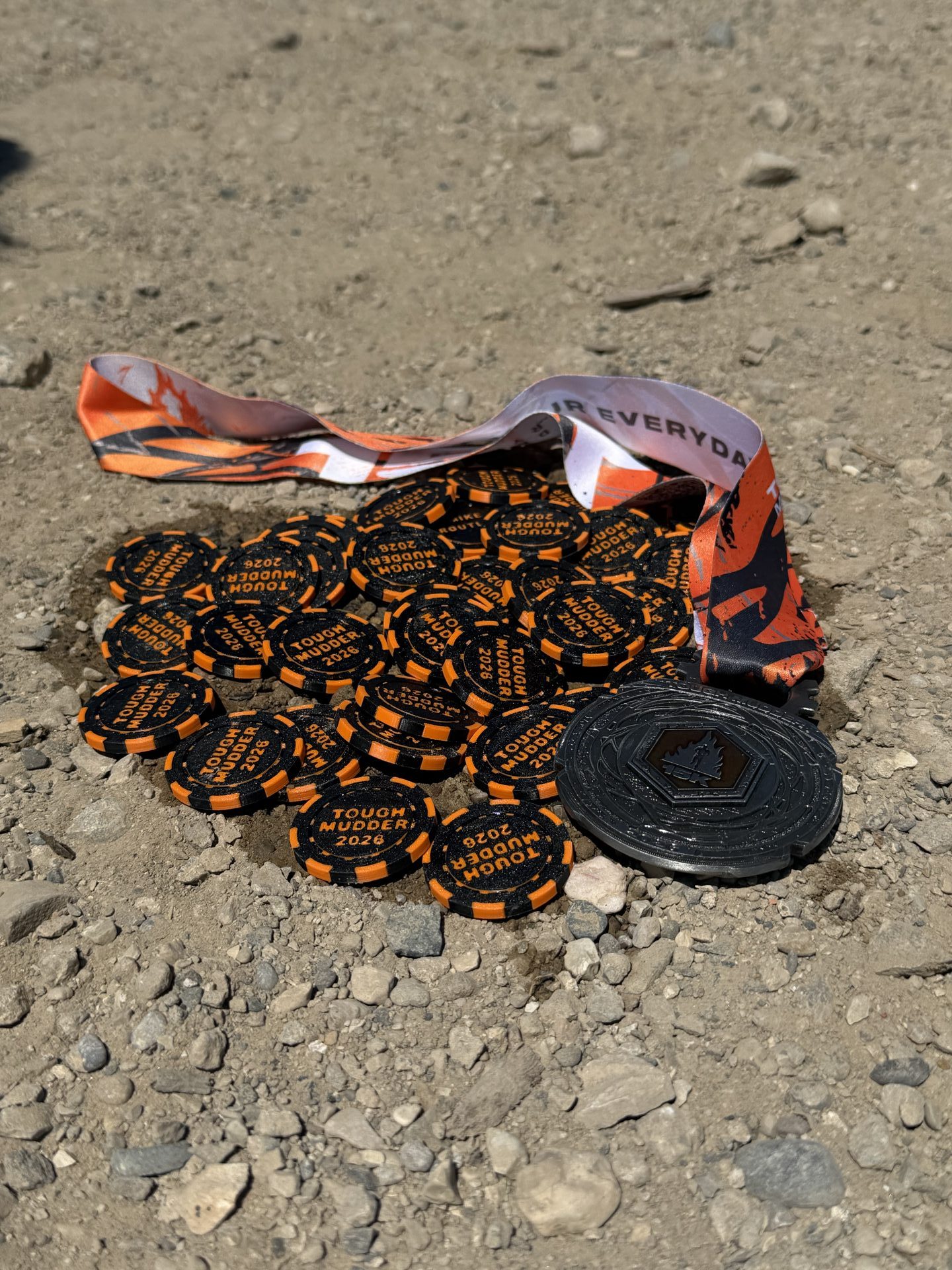 27 Tough Mudder honor coins laid out with the finisher medal in the dirt
